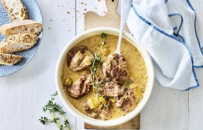 Een heerlijke portie varkenswangetjes is geserveerd in een groot wit bord, de saus heeft een goudkleurige kleur. Hij is gemaakt op basis van mosterd, room en Vermouth. Het gerecht is afgewerkt met enkele takjes verse tijm en er staat een licht blauw bordje langs met stukjes stokbrood. 