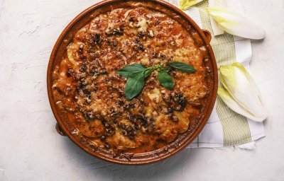 Op het aanrecht staat een ronde ovenschaal die net uit de oven komt. Hij heeft een krokant kaaskorstje en enkele vers witloofblaadjes rond hem. De ovenschotel is er nl eentje van witloof, aardappel, spek en tomatensaus.
