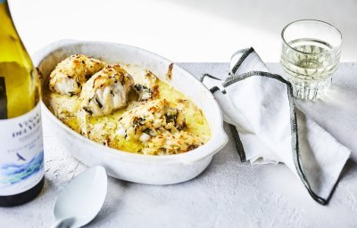 Een ovenschotel staat klaar op tafel om meteen te beginnen. De schotel is gevuld met stukken zeeduivel en prei in een mosterdsausje. Er ligt een krokant korstje op van geraspte aardappel en kaas. Er staat een flesje wijn langs en een witte scheplepel.