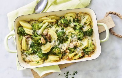 Een ovenschotel van aardappel, broccoli, champignons en blauwe kaas staat op het midden van het marmere aanrecht op een houten snijplank. Verse kruiden zorgen voor een extra groene touch!