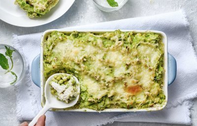 Eten is klaar! De ovenschotel staat op een witte dikke doek en een porseleine lepel schept de eerste portie uit de schotel. De parmentier heeft een mooie frisse groene kleur door de vele erwtjes. 