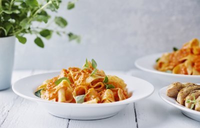 Een portie pasta met homemade paprikasaus staat klaar op een witte houten tafel. Een vers plantje oregano staat achteraan in beeld. 