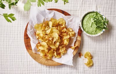 Op tafel ligt een kalkwit tafelkleed, met hierop een houten bordje met wit boterpapiertje en chips van pastinaak op. Er staat een dippotje langs met de dip van broccoli en roomkaas. 