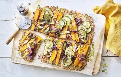 Een krokante bodem van bloemkool en walnoten waarop kleurrijke groentjes liggen zoals wortel, courgetteplakjes en rode ui. De plaattaart ligt op een houten snijplank waar een pizzasnijder langs ligt, de pepermolen en goudgele servette. 