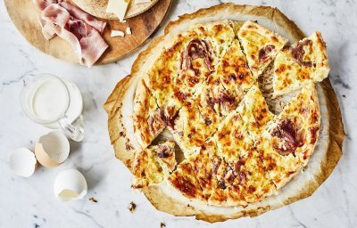 Vers uit de oven staat deze quiche te schitteren op het marmere aanrecht. Hij is aangesneden in kleinere stukjes om aan te vallen tijdens het aperomoment. Er liggen enkele eitjes naast ter decoratie en verse boerenham.