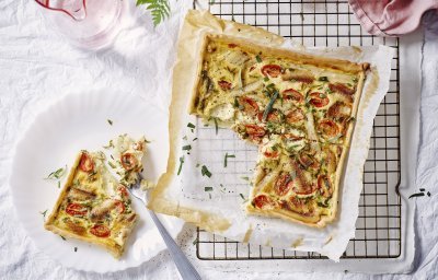 Vers gebakken uit de oven staat de quiche met asperges, tomaten en gerookte paling te blinken op tafel. Er werd al een stukje aangesneden en op een bordje gepresenteerd. Roze linnen servet en waterkaraf zorgen voor de sfeer in de foto. 