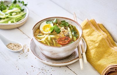 Een goeie portie soep staat uitgeschept op een houten witte tafel. De Japanse soep bestaat uit noedels van courgette aangevuld met peultjes, puntpaprika en kip. De soep is afgewerkt met een rood schijfje van een chilipeper en een gekookt eitje. 