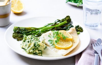 Op tafel staat een wit groot bord met hierop de roggevleugel, citroenschijfjes, gegrilde bimi en een schep kervelpuree. Er staat een glas water langs, samen met een roze linnen servette en bestek.