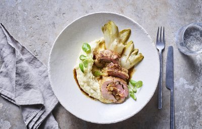 Een stukje rollade van varkensvlees ligt in een wit groot bordje op een smeuige pastinaakpuree met gebakken witloofstronkjes. 