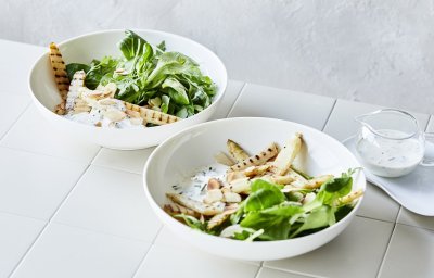 Salade met gegrilde asperges in 2 diepe borden. De groene salade bestaat uit veldsla en rucola met daarbovenop gegrilde witte asperges. Er is ook een yoghurtdressing met verse kruiden die bovenaan het bord staat in een glazen karafje. 