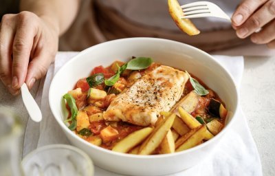 Aan taffel zit een vrouw die een stukje aardappel prikte op haar vork. Voor haar staat een bordje gevuld met het gerecht, vis met Prvençaalse saus en gebakken aardappelpartjes. 