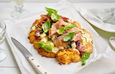 Een aardappelpizza ligt klaar om aan te snijden, en wat voor. Het is er eentje met een bodem van smashed potatoes, belegd met zure room, coutgette en rode ui. En afgewerkt met gedroogde ham en basilicumblaadjes. 