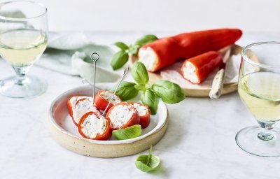 Een opgevulde puntpaprika met roomkaas en kruiden ligt op een houten snijplankje. Er werden rolletjes van gesneden en vastgeprikt met een stokje om het aperitief in stijl te starten. Voor de garnering en de kleur voegde we nog enkele verse blaadjes basilicum toe.