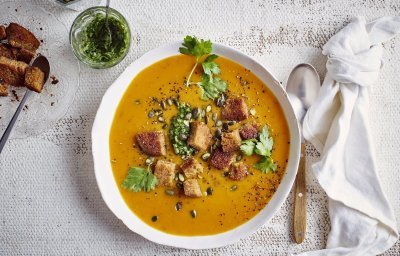Op tafel staat een wit soepbord gevuld met heerlijk oranje pompoensoep. De soep is afgewerkt met verse groene kruiden, pompoenpitten en croutons van zuurdesembrood. Er ligt een witte linnen servette langs, geknoopt, en een soeplepel. 