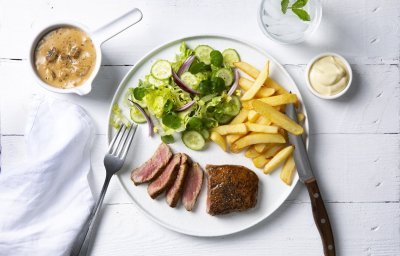 Een echte Vlaamse klassieker die smaakt voor jong & oud, een steak mooi gebakken met heerlijke goude frietjes en een salade. De champignonsaus staat langs het bord in een sauspotje. Bestek en servette liggen langs het bord klaar om te starten. 