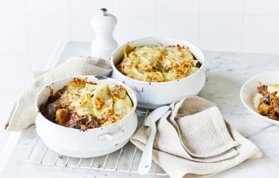 Twee witte stoofpotjes sttaan op een witte houten tafel en zijn gevuld met traag gegaard rundsvlees. De stoofpot is afgedekt met een laagje van aardappelschijfjes. De gratin is mooi goudgeel gebakken. Er staat een pepermolen langsheen de kookpotten, servette en grote lepel.