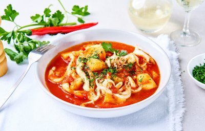 Een tomatenrood stoofpotje met vis staat op tafel. Het is een stoofpotje met stukjes paprika, aardappelen en tomatensaus. 