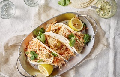 Drie wraps liggen gepresenteerd in een ovenschotel van inox. Deze staat op een gedekte tafel met wit linnen kleed. Er liggen twee citroenhelften bij de tortilla's en verse koriander. 