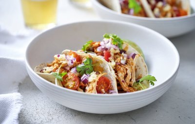 In een diep wit bord liggen twee gevulde taco's met pulled chicken, afgewerkt met tomaat, rode ui en geraspte Passendael kaas. Om wat extra kleur te geven liggen er enkele blaadjes koriander op. Wij serveren ze met frisgekoeld biertje, smakelijk!