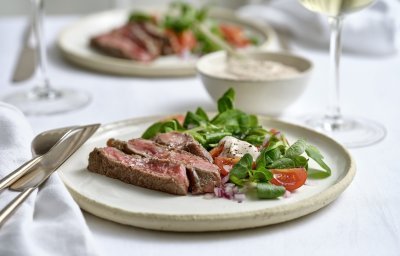 Twee borden zijn geserveerd op tafel, de licht beige borden zijn gevuld met reepjes gegrild vlees op een gemengde en frisse sla van veldsla en kerstomaten. Er staat ook een glaasje witte wijn op tafel.  