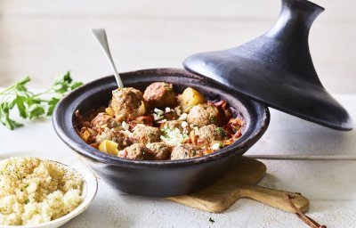 Zwartkleurige tajine met gehaktballetjes van lamsvlees, afgewerkt met verbrokkelde feta. Er staat een portie couscous naast de tajine. 