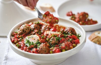 Op tafel staat een witte tajine heerlijk gevuld met gehaktballen in een rijke tomatensaus met eitjes. Er wordt net een portie uitgeschept. 