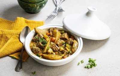 Op tafel staat een witte tajine met een gerechtje van rundvlees, courgette, selder, wortel en stukjes aardappel. Er ligt een okerkleurige servette bij en groene kommetjes om de tajine in op te scheppen. 