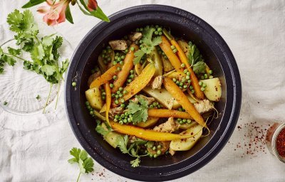 Een zwarte tajine waarin je kleurrijke ingrediënten vind. Denk maar aan wortel, aardappel, erwtjes en vlees. Er liggen nog wat verse kruiden naast om en een bloemetje voor de juiste sfeer in het beeld te krijgen. 