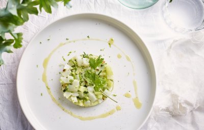 Op tafel ligt een mooi wit linnen tafelkleed, waarop het witte bord staat. Hierop ligt de tartaar van zeekat met komkommerstukjes, lente-ui en koriander. We voegden ook wat chili toe voor extra pit en als afwerking nog wat olijfolie en een korianderblaadje. 