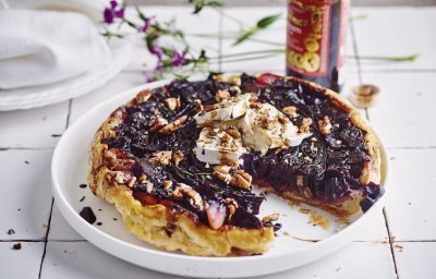 Een tarte tatin van rode kool met geitenkaas ligt op een groot wit bord. Er is al een stukje aangesneden en ze werd afgewerkt met enkele druppels balsamico en pecannoten. Achteraan zie je de fles balsamico nog op het aanrecht staan alsook een paars bloemetje. 