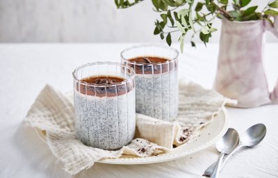 Op een bord staan twee grote glazen gevuld met de chiapudding, hij is afgewerkt bovenaan met cacaopoeder en chocoladeschilfers. Er liggen twee zilvere lepeltjes langs.