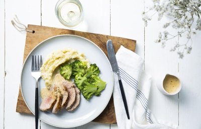 Op tafel ligt een houten snijplank met hierop een lichtblauw groot plat bord. Hierop is het hoofdgerecht geserveerd, stukjes varkenshaasje in sneetjes, plakjes broccoli en een een schep aardappelpuree. Het sausje zit in een sauskannetje langsheen het bord.