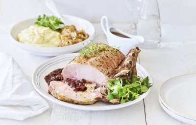 Een varkenskroontje ligt op een groot wit bord, aangesneden en opgediend met de rode wijn saus. In de achtergrond staat de aardappelpuree en de stukjes witloof. Het varkensvlees is mooi roos gebakken. 