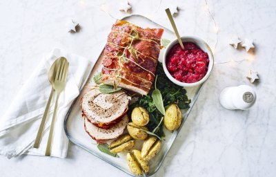 Een mooi aangesneden varkensrollade ligt op een rechthoekig dienblad. De aardappeltjes werden net niet doormidden gesneden alvorens ze de oven in gingen met een kruidentakje. De veenbessencompote staat langs de rollade die zorgt voor een intense kleur in deze feestmaaltijd. Goud bestek ligt langs het gerecht. 