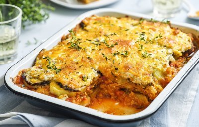 Er staat een grote schotel met vegetarische moussaka op tafel. Er is al een portie uitgeschept en op een bordje geserveerd. De moussaka is afgewerkt met verse tijmblaadjes. Op tafel staan ook nog 2 glazen en het plantje verse tijm.