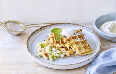 Wafeltjes van verloren brood liggen op een licht grijs bordje. De stukjes fruit van appel en peer liggen onder de wafeltjes. We gaven ook een toefje slagroom op de wafels voor extra smaak. Er ligt een blauwe servette en klein zeefje met bloemsuiker langs het bord.