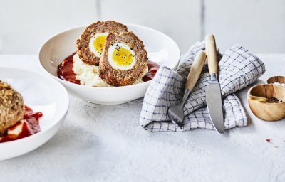 In een diep wit bord ligt de aardappelpuree onderaan met verse rode tomatensaus en daar bovenop de doormidden gesneden vogelnestjes. Er ligt een handdoek langs met blauwe ruitjes, hierop het bestek met houten handvat. 