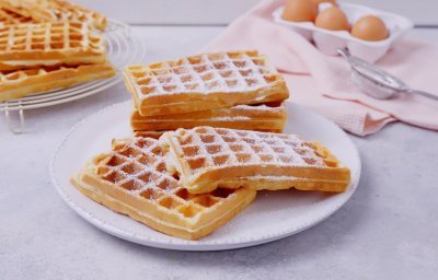 Een stappeltje klaargemaakte wafels ligt op een groot wit bord. Bloemsuiker is rijkelijk op de wafels gestrooid. In de achtergrond zie je nog enkele eitjes liggen en een roze linnen servette.