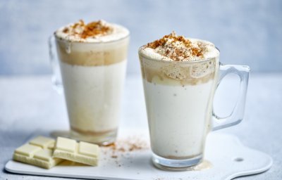 Twee hoge glazen tassen staan op het aanrecht klaar om te serveren. Ze zijn gevuld met witte chocolademelk, een goeie toef speculaasroom er bovenop en wat speculaaskruim. Er ligt een stukje witte chocolade naast. 