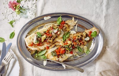 Twee filets van zeetong liggen op een inoxen serveerschaal en staan op de gedekte tafel. Op de vis ligt een fijngesneden pesto van zuiderse groenten. 