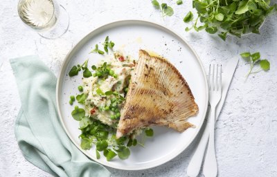 Een wit bord gevuld met een frisse schep van de puree boordevol extra groentjes, denk maar aan verse lente-uitjes, zongedroogde tomaatjes, waterkers, kruiden enz. Hierop werd de gebakken rogvleugel gelegd die mooi krokant is gebakken. Naast het bord ligt ook nog een lichtblauwe servette, kommetje met extra waterkers en een glaasje wijn. Smakelijk!