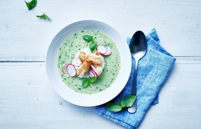 Wit diep bord met gazpacho van komkommer, tongscharreepjes, radijsjes en basilicum. Rechts naast het bord ligt een lepel op een blauw servette.