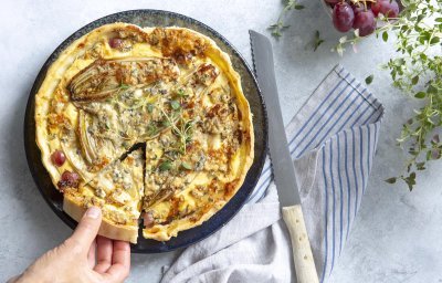 Quiche met witloof, blauwe kaas en druiven. De herfst gecombineerd met fruit. De quiche is al aangesneden en een hand neemt een taartstuk uit de quiche. Verse tijm zorgt voor sfeer en staat rechtsbovenaan in de foto.