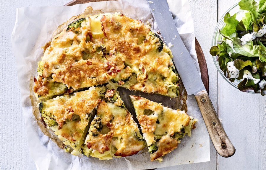 Deze hartige taart lijkt op een quiche maar is toch anders. Deze is gemaakt van aardappelschijfjes, broccoli en kippenwit. Ze ligt op een houten ronde snijplank met boterpapiertje onder, ze is al voor de helft aangesneden. Rechtsboven staat nog een kommetje gemengde sla. 