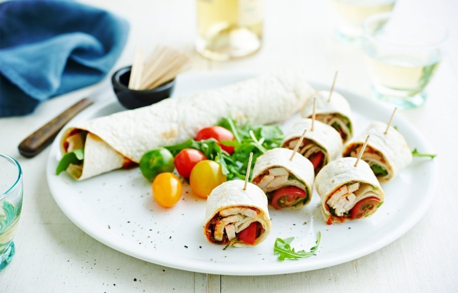 Op een bord ligt een rol wrap met groenten, wat kerstomaten en zes stukken wrap met een prikker in.