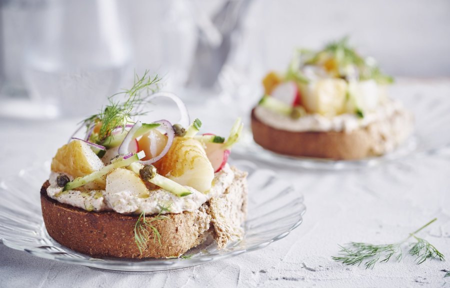 Een goedgevulde bruschetta ligt op een klein glazen bordje en staat op tafel. De bruschetta werd afgewerkt met een takje dille. Achteraan de tafel staat een kan met water en bestek om deze toast te verorberen. 