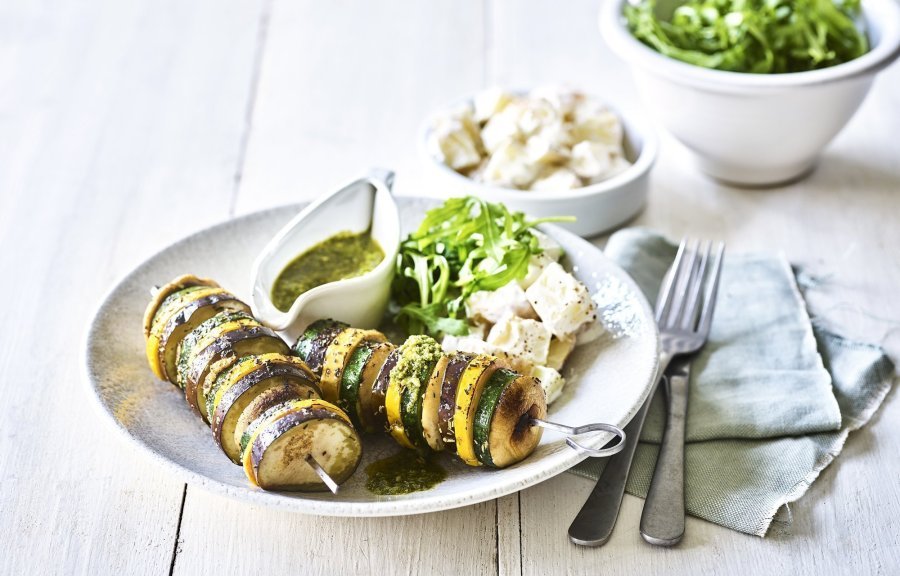 Op een bleke houten tafel staat een diep bord met hierin een schep aardappelsalade in, twee groentespiesjes met schijfjes van gegrilde aubergine, courgette, champignons en een kannetje met rucolapesto. Verder staat er ook nog een kom met verse groene salade, bestek en een muntkleurige servette.