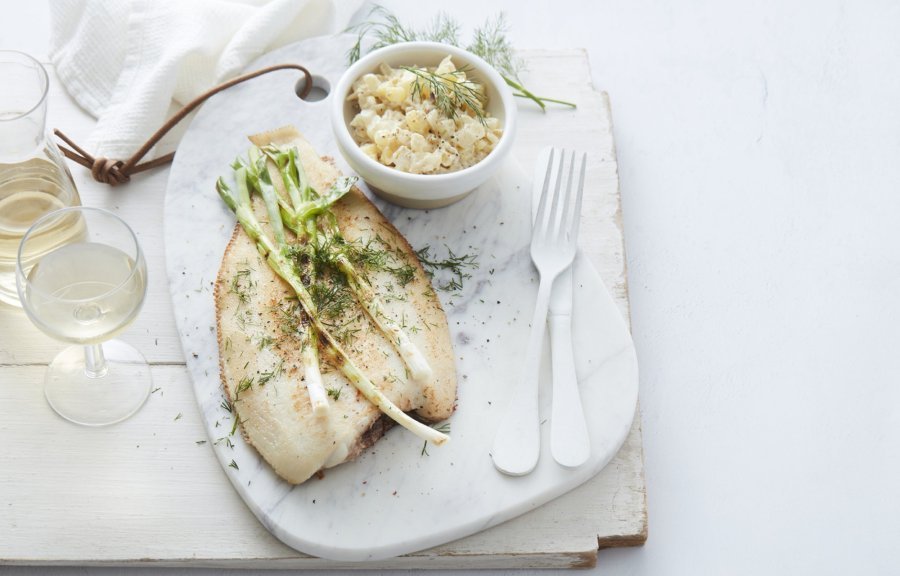 De gebakken vis ligt op een stenen witte snijplank, waar de gigrilde lente-ui bovenop ligt. De Macédoine van venkel en krieltjes is opgediend in een apart wit potje. Er ligt wit bestek langs, met een glaasje witte wijn en een linnen servette. Het gerechtje is afgewerkt met verse dille.