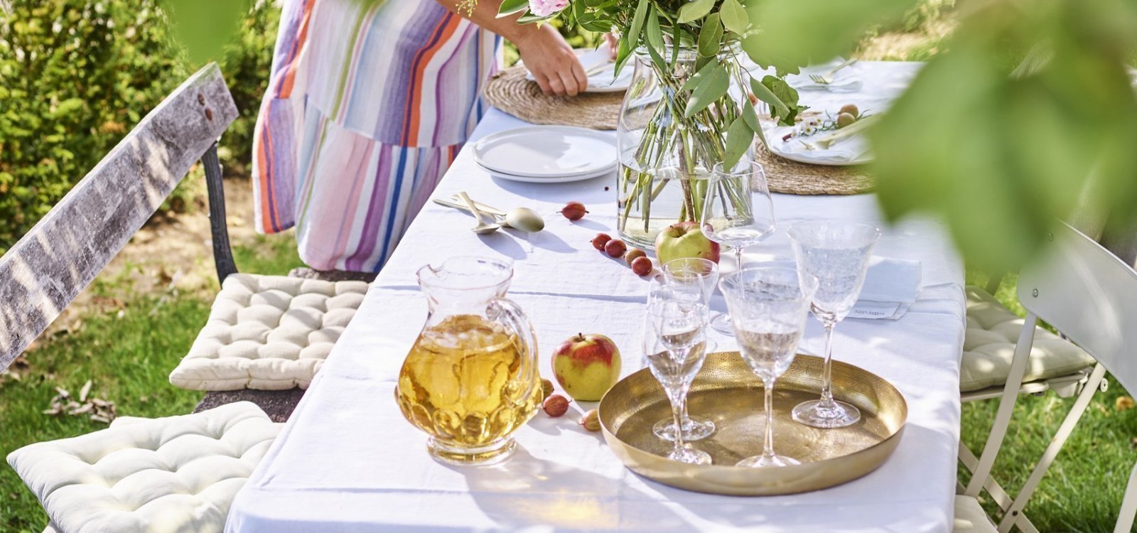 Vrouw met gestreept zomerjurk is de tafel aan klaarzetten voor enkele gasten. Er staat al een kan met vers appelsap op tafel en enkele glazen. De vrouw is de borden aan het plaatsen op de tafel met wit linnen tafelkleed. De tafel staat onder een perenboom in de tuin. 