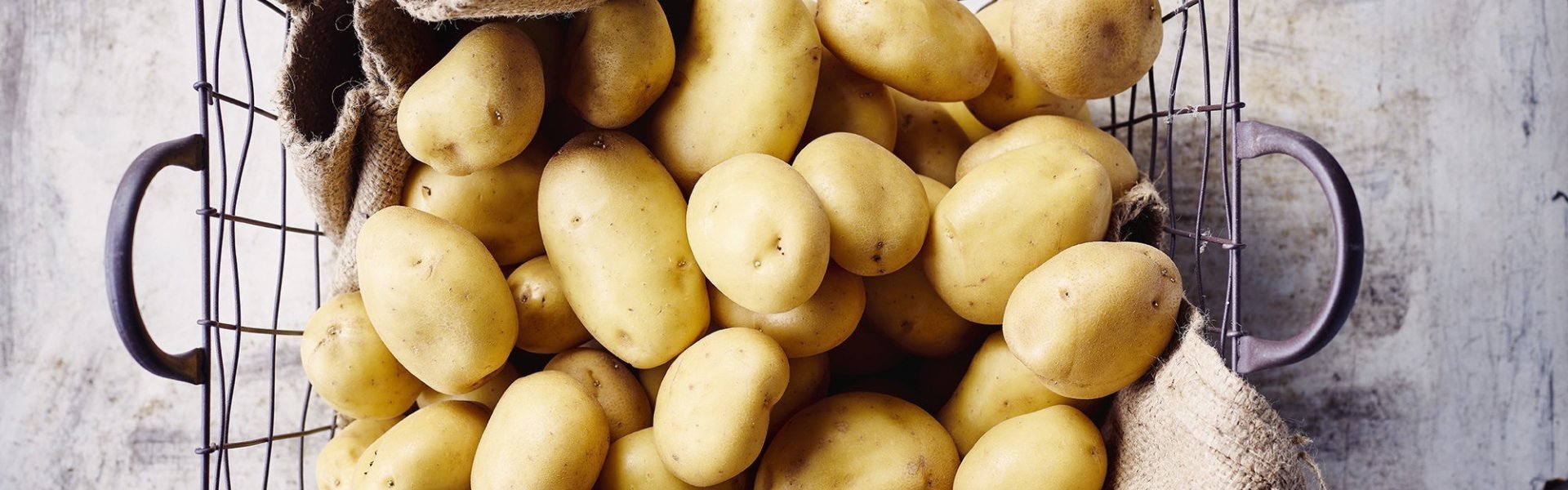 Aardappelen Aardappelen liggen in ijzeren mand met bruine linnen doek onder. De mand staat op een grijze gietbeton.